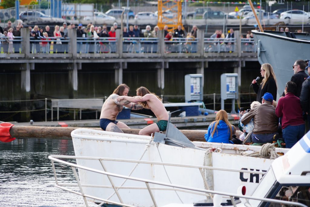 Two men try to push each other off a log as people watch. 