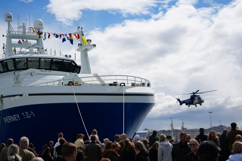 A crowd at a harbor watches as a coast guard helicopter hovers beside a ship.