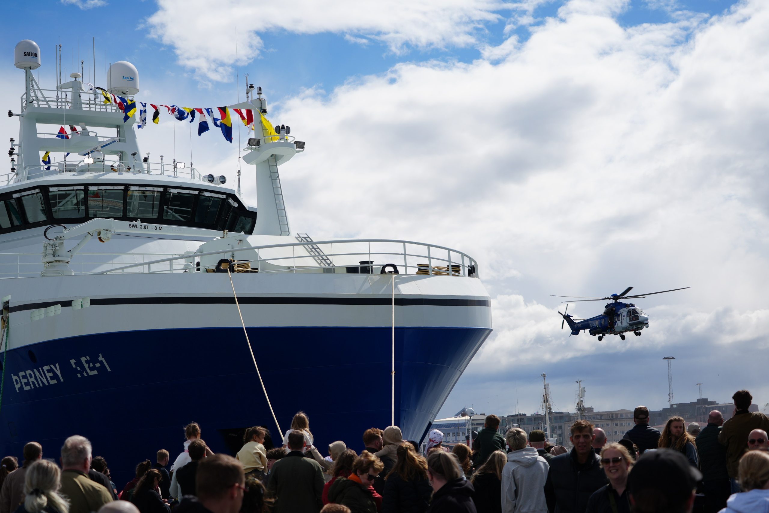 A crowd at a harbor watches as a coast guard helicopter hovers beside a ship.