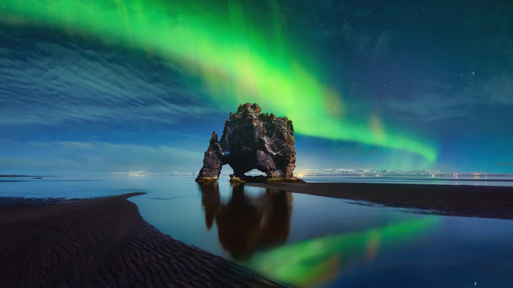 Northern lights glow above Hvítserkur sea stack in northern Iceland, reflected in the water.