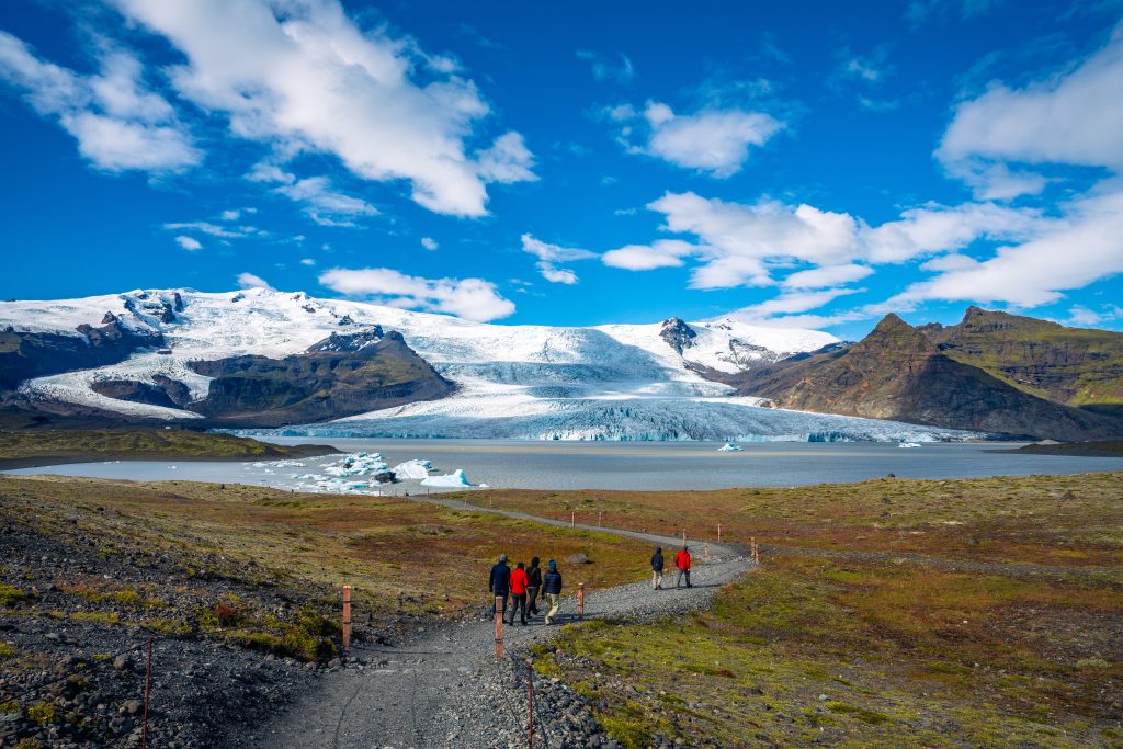 MICE in Iceland – A glacier flowing between rugged mountains into a calm glacial lagoon, with small icebergs and hikers walking along a gravel path beneath a vivid blue sky.