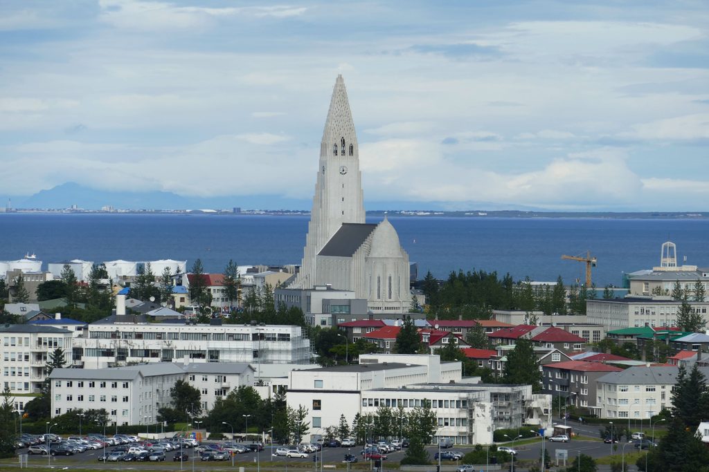 Hallgrímskirkja church rising above central Reykjavik, with surrounding buildings, red rooftops, and the Atlantic Ocean in the background.