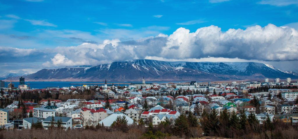 Central Reykjavik, Iceland, with colorful rooftops overlooking the waterfront and snow-streaked Mount Esja beneath dramatic clouds and a bright blue sky.
